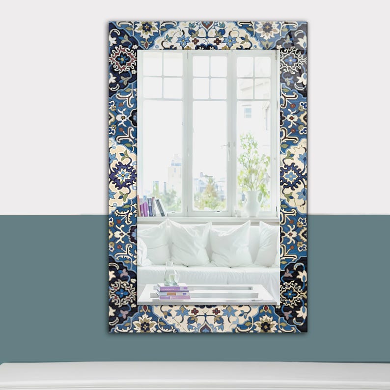 K&ouml;nnte beinhalten: Rechteckiger Spiegel mit einem dekorativen Rand mit blauem, wei&szlig;em und beigem Blumenmuster. Der Spiegel reflektiert ein helles Fenster mit wei&szlig;em Rahmen und einen Blick auf einen Raum mit wei&szlig;en Kissen und einem wei&szlig;en Sofa. Der Spiegel ist an einer zweifarbigen Wand befestigt.