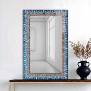 Può includere: Specchio rettangolare con bordo decorativo a motivi blu e oro. Lo specchio è esposto su una mensola marrone scuro. Un vaso nero con piante secche è a destra. Lo specchio riflette una stanza bianca con una finestra.