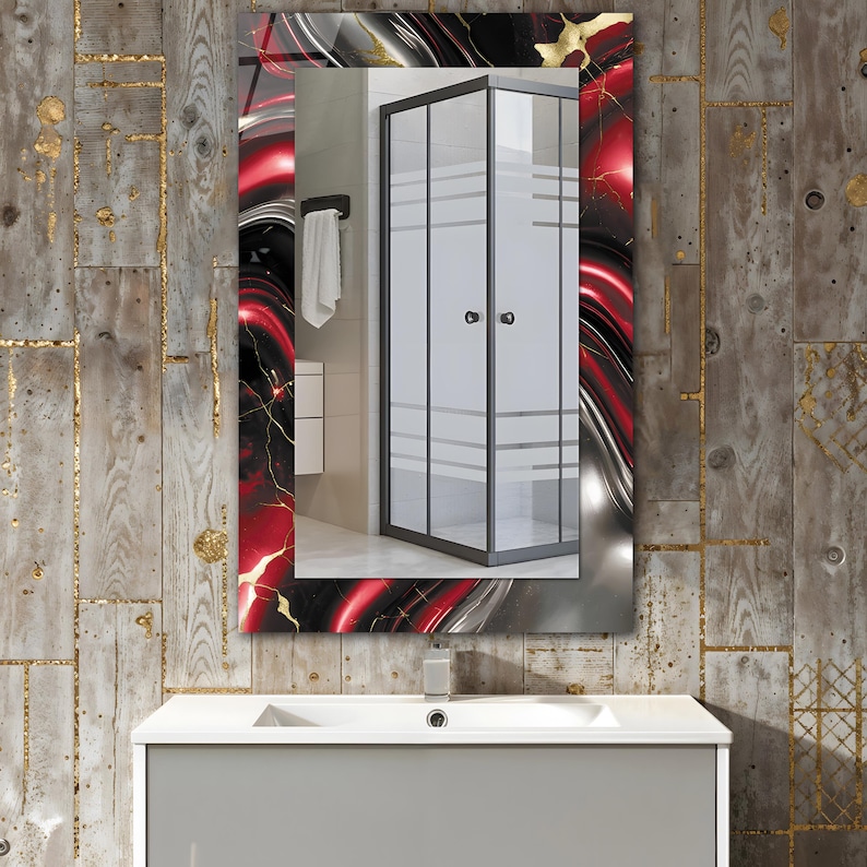 May include: Rectangular bathroom mirror with a decorative frame featuring red, black, and gold abstract patterns. The mirror reflects a shower enclosure with frosted glass doors and a white towel. The mirror is mounted above a gray vanity.