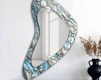 May include: An abstract-shaped wall mirror with a mosaic-style border. The border features a blend of blue, white, and gray tones, with circular and flowing patterns. The mirror is displayed on a dark wooden shelf against a white wall.