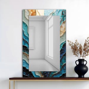 Puede incluir: Un espejo de pared rectangular con un borde decorativo. El borde presenta un patrón ondulado en tonos de azul, turquesa, dorado y blanco. El espejo refleja una habitación con una ventana y una pared blanca. Un jarrón negro con plantas secas está sobre una repisa de madera.