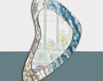 May include: A decorative wall mirror with a unique, asymmetrical shape. The frame features a mosaic pattern in shades of blue, white, and beige, with gold accents. The mirror reflects a bright room with a window and white furniture.