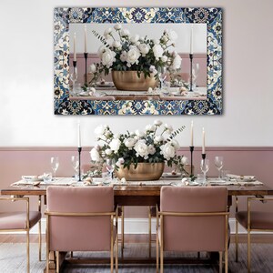 K&ouml;nnte beinhalten: Ein rechteckiger Spiegel mit einem dekorativen blau-wei&szlig;en Musterrahmen h&auml;ngt &uuml;ber einem Esstisch. Der Tisch ist mit einer goldenen Schale mit wei&szlig;en Blumen, Kerzen und Gedecken gedeckt. Rosafarbene St&uuml;hle mit goldenen Rahmen umgeben den Tisch.