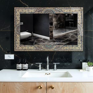 K&ouml;nnte beinhalten: Rechteckiger Spiegel mit einem verzierten, gemusterten Rahmen in Gold-, Blau- und Beiget&ouml;nen. Der Spiegel spiegelt ein Badezimmer mit einem wei&szlig;en Waschbecken, einer Chromarmatur und schwarzen Marmorw&auml;nden wider. Der Waschtisch ist aus hellem Holz.