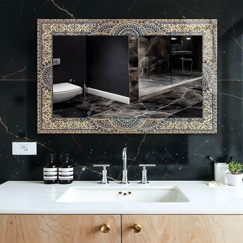 K&ouml;nnte beinhalten: Rechteckiger Spiegel mit einem verzierten, gemusterten Rahmen in Gold-, Blau- und Beiget&ouml;nen. Der Spiegel spiegelt ein Badezimmer mit einem wei&szlig;en Waschbecken, einer Chromarmatur und schwarzen Marmorw&auml;nden wider. Der Waschtisch ist aus hellem Holz.