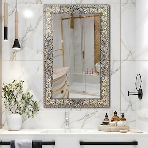 K&ouml;nnte beinhalten: Rechteckiger Badezimmerspiegel mit einem verzierten, gemusterten Rand in Gold, Blau und Creme. Der Spiegel spiegelt ein Badezimmerinterieur mit einem wei&szlig;en Waschbecken, Marmorw&auml;nden und einer Dusche wider. Ein kleiner schwarzer, runder Spiegel ist an der Wand befestigt.