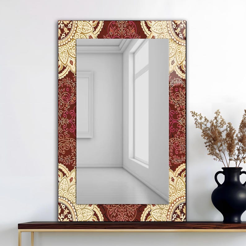 May include: Rectangular mirror with a decorative border featuring a repeating pattern in burgundy, cream, and pink. The mirror reflects a white room with a window and a framed picture. A black vase with dried plants sits on a wooden shelf.