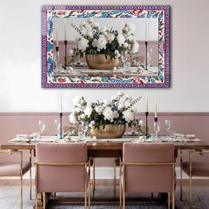 May include: A rectangular mirror with a decorative border featuring a floral and geometric pattern in red, blue, and white. The mirror reflects a dining table set with a floral centerpiece and candles.