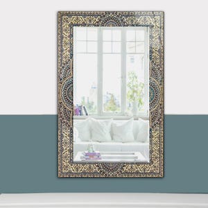 K&ouml;nnte beinhalten: Rechteckiger Wandspiegel mit einem verzierten, gemusterten Rahmen in Beige-, Braun- und T&uuml;rkist&ouml;nen. Der Spiegel reflektiert einen Raum mit einem wei&szlig;en Sofa, Kissen und einem Fenster mit Blick auf eine Stadtlandschaft. Der Rahmen hat ein detailliertes, traditionelles Design.