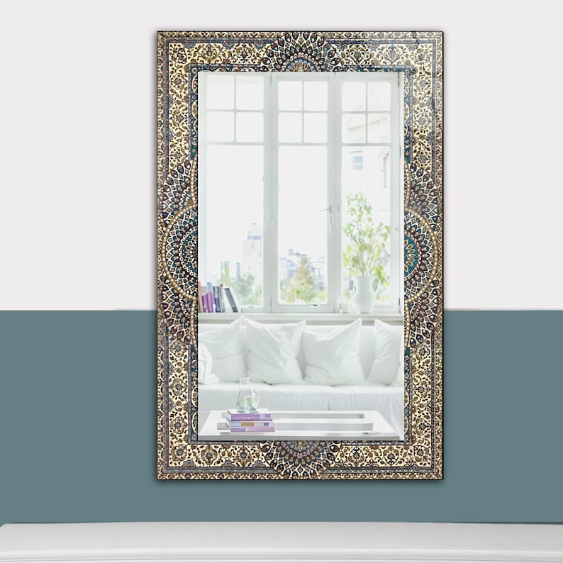K&ouml;nnte beinhalten: Rechteckiger Wandspiegel mit einem verzierten, gemusterten Rahmen in Beige-, Braun- und T&uuml;rkist&ouml;nen. Der Spiegel reflektiert einen Raum mit einem wei&szlig;en Sofa, Kissen und einem Fenster mit Blick auf eine Stadtlandschaft. Der Rahmen hat ein detailliertes, traditionelles Design.
