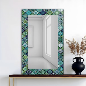 May include: Rectangular mirror with a decorative border featuring a repeating pattern of blue, green, and white geometric shapes. The mirror is displayed on a dark wooden shelf, with a black vase and dried plants to the right.