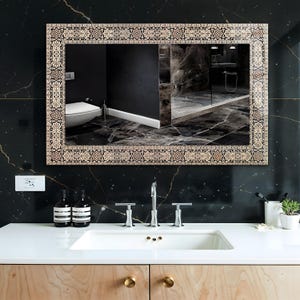 May include: Rectangular mirror with a decorative border featuring a repeating pattern in beige, brown, and black. The mirror is mounted on a black marble wall above a white sink with chrome fixtures. The reflection shows a bathroom.