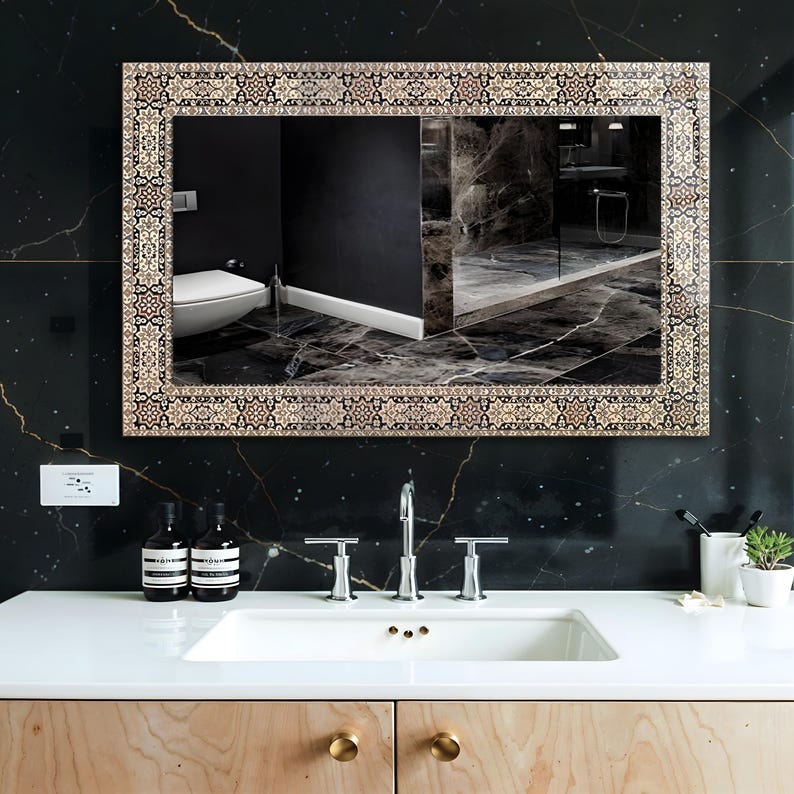 May include: Rectangular mirror with a decorative border featuring a repeating pattern in beige, brown, and black. The mirror is mounted on a black marble wall above a white sink with chrome fixtures. The reflection shows a bathroom.