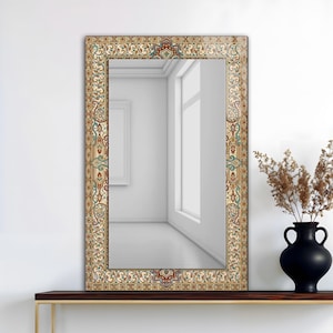 Puede incluir: Espejo rectangular con un marco decorativo con un patrón detallado y colorido en tonos beige, rojo y turquesa. El espejo se exhibe sobre una estantería marrón oscuro, con un jarrón negro que contiene plantas secas a la derecha.