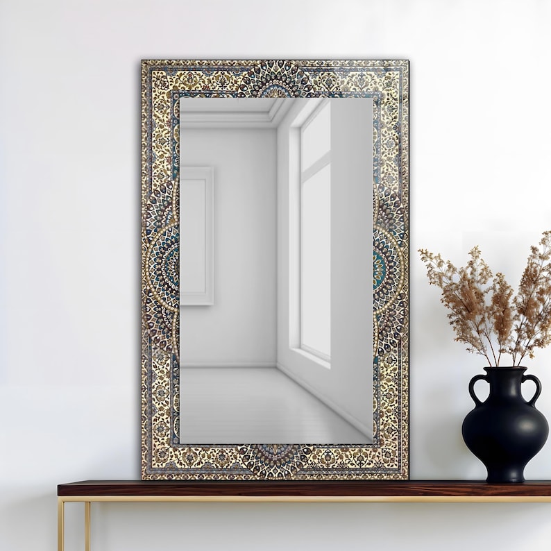 K&ouml;nnte beinhalten: Rechteckiger Spiegel mit einem verzierten, gemusterten Rahmen in Beige-, Blau- und Goldt&ouml;nen. Der Spiegel reflektiert einen hellen Raum mit einem Fenster und einem gerahmten Bild. Eine schwarze Vase mit getrockneten Pflanzen steht auf einem Holzregal.