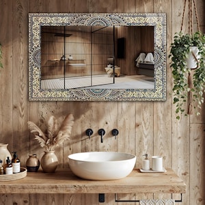 K&ouml;nnte beinhalten: Ein rechteckiger Spiegel mit einem verzierten, gemusterten Rahmen h&auml;ngt &uuml;ber einem h&ouml;lzernen Waschtisch. Der Spiegel spiegelt ein Badezimmer mit Dusche und Holzw&auml;nden wider. Ein wei&szlig;es Keramikwaschbecken und schwarze Wasserh&auml;hne sind sichtbar.