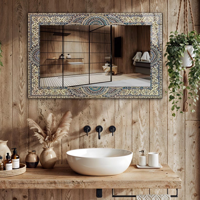 K&ouml;nnte beinhalten: Ein rechteckiger Spiegel mit einem verzierten, gemusterten Rahmen h&auml;ngt &uuml;ber einem h&ouml;lzernen Waschtisch. Der Spiegel spiegelt ein Badezimmer mit Dusche und Holzw&auml;nden wider. Ein wei&szlig;es Keramikwaschbecken und schwarze Wasserh&auml;hne sind sichtbar.