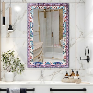 May include: Rectangular mirror with a decorative border featuring a floral pattern in red, blue, and white. The mirror is mounted on a white tiled wall above a bathroom sink. The mirror reflects a bathroom interior.