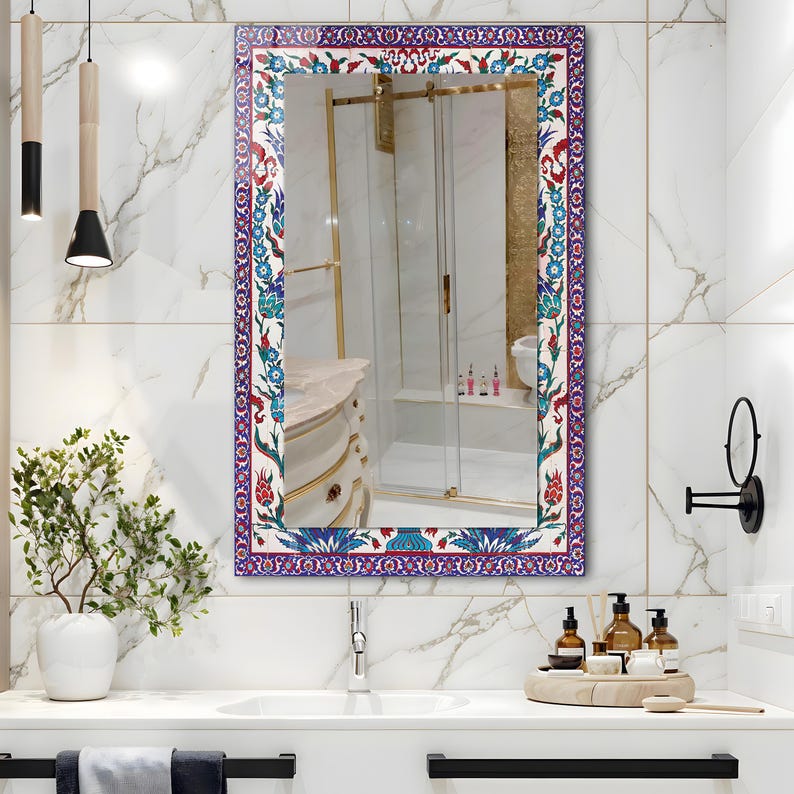 May include: Rectangular mirror with a decorative border featuring a floral pattern in red, blue, and white. The mirror is mounted on a white tiled wall above a bathroom sink. The mirror reflects a bathroom interior.