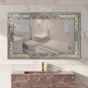 K&ouml;nnte beinhalten: Rechteckiger Badezimmerspiegel mit einem verzierten, gemusterten Rand in Blau-, Gold- und Cremet&ouml;nen. Der Spiegel spiegelt ein modernes Badezimmer mit wei&szlig;er Toilette, Dusche und Waschtisch wider. Unter dem Spiegel ist ein goldener Wasserhahn zu sehen.