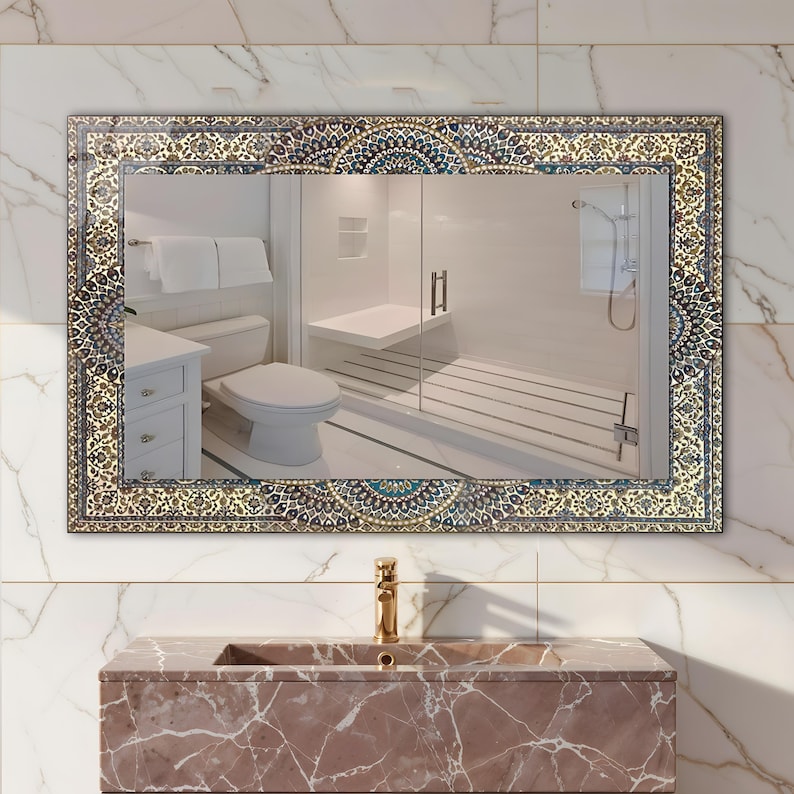 K&ouml;nnte beinhalten: Rechteckiger Badezimmerspiegel mit einem verzierten, gemusterten Rand in Blau-, Gold- und Cremet&ouml;nen. Der Spiegel spiegelt ein modernes Badezimmer mit wei&szlig;er Toilette, Dusche und Waschtisch wider. Unter dem Spiegel ist ein goldener Wasserhahn zu sehen.