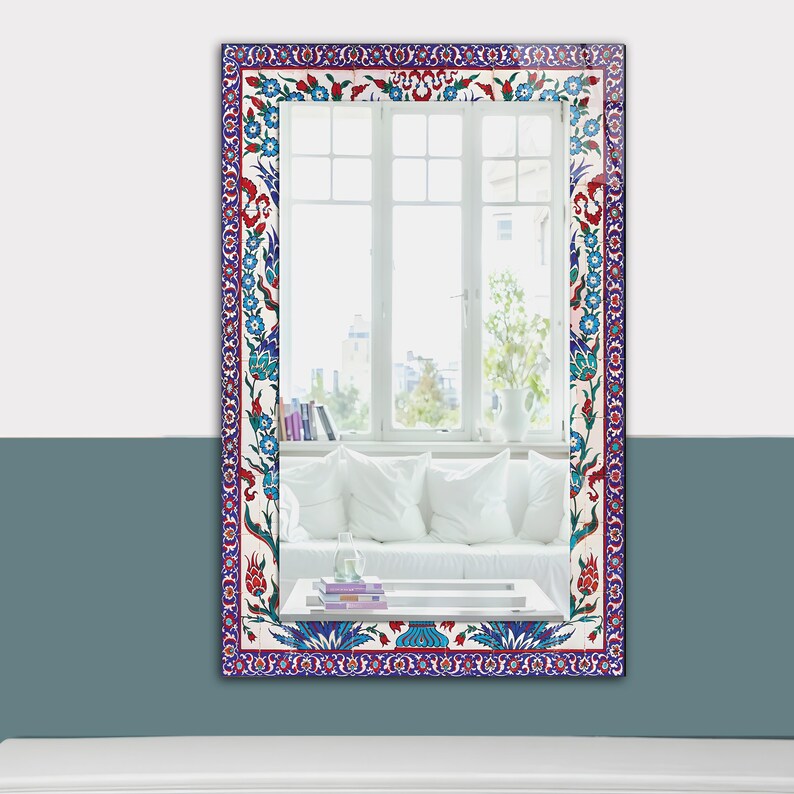 May include: Rectangular mirror with a decorative border featuring a floral pattern in red, blue, and white. The mirror reflects a bright room with a window, white sofa, and books. The frame's design is reminiscent of traditional tilework.