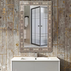 May include: Rectangular bathroom mirror with an ornate, patterned border in shades of brown, beige, and black. The mirror reflects a shower enclosure with frosted glass panels and a white towel. The mirror is mounted above a gray vanity.