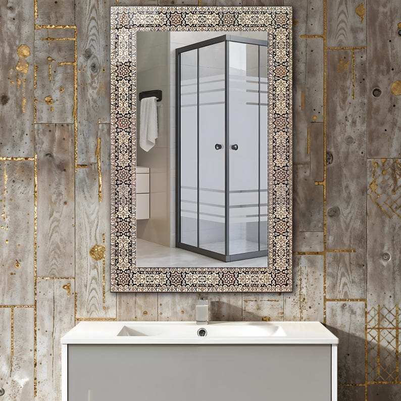 May include: Rectangular bathroom mirror with an ornate, patterned border in shades of brown, beige, and black. The mirror reflects a shower enclosure with frosted glass panels and a white towel. The mirror is mounted above a gray vanity.