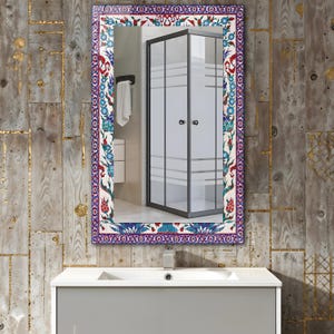 May include: Rectangular mirror with a decorative border featuring a floral pattern in red, blue, and white. The mirror reflects a bathroom scene with a shower enclosure and a white sink.