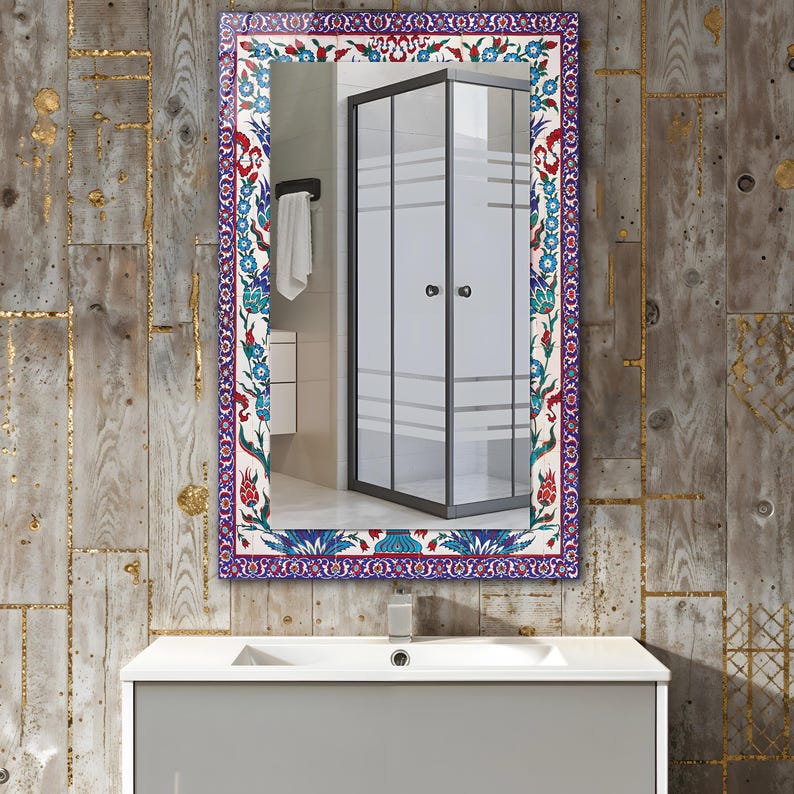 May include: Rectangular mirror with a decorative border featuring a floral pattern in red, blue, and white. The mirror reflects a bathroom scene with a shower enclosure and a white sink.