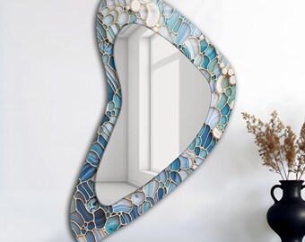 May include: A decorative wall mirror with a unique, asymmetrical shape. The frame features a mosaic design with shades of blue, white, and gold. The mirror is displayed on a dark wooden shelf, with a black vase and dried plants in the background.