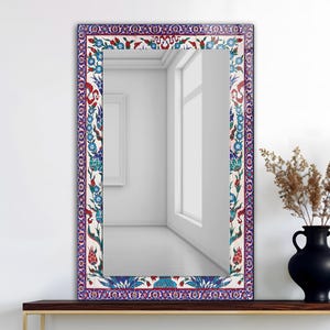 May include: Rectangular mirror with a decorative border featuring a floral pattern in red, blue, and white. The mirror is displayed on a dark wooden surface, with a black vase and dried plants visible in the background.