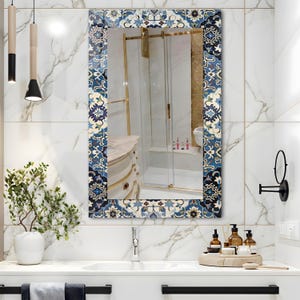 K&ouml;nnte beinhalten: Rechteckiger Badezimmerspiegel mit dekorativem Rand mit blau-wei&szlig;em Blumenfliesenmuster. Der Spiegel spiegelt ein Badezimmerinterieur mit wei&szlig;em Waschbecken, Marmorw&auml;nden und goldenen Akzenten wider.