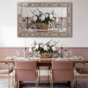 May include: A dining room scene featuring a large mirror with a decorative border above a wooden table set for a meal. The table is adorned with a floral centerpiece in a gold bowl, candles, and place settings. Pink chairs with gold frames complete the elegant setting.