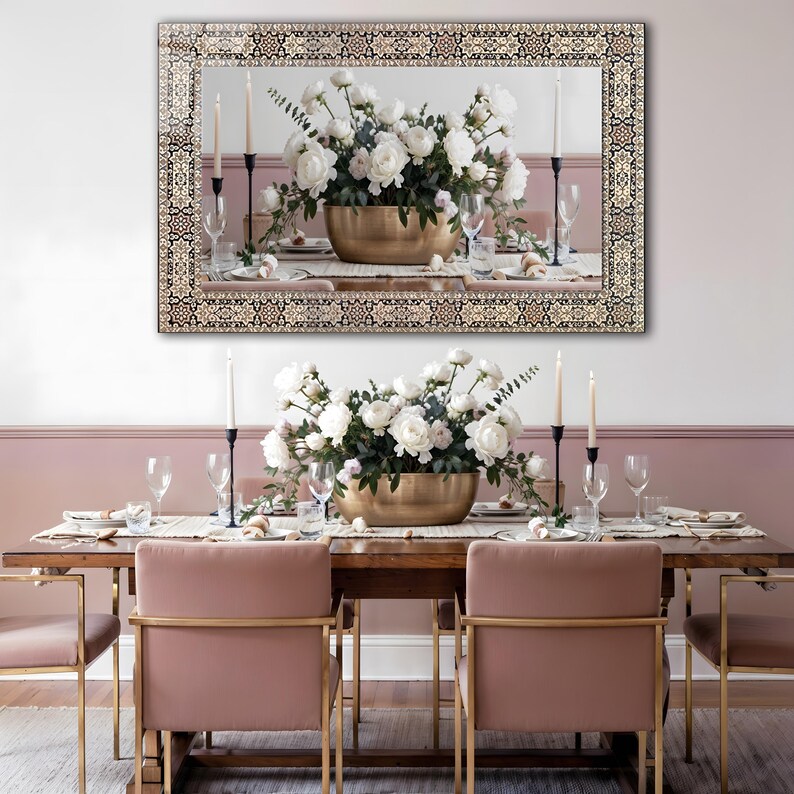 May include: A dining room scene featuring a large mirror with a decorative border above a wooden table set for a meal. The table is adorned with a floral centerpiece in a gold bowl, candles, and place settings. Pink chairs with gold frames complete the elegant setting.