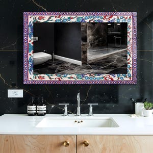 May include: Rectangular mirror with a decorative floral border in red, blue, and white. The mirror is mounted on a black marble wall above a white sink. The bathroom features a modern design with wooden cabinets and gold hardware.