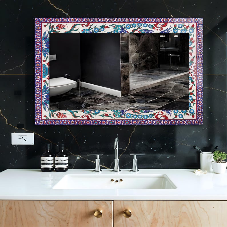 May include: Rectangular mirror with a decorative floral border in red, blue, and white. The mirror is mounted on a black marble wall above a white sink. The bathroom features a modern design with wooden cabinets and gold hardware.