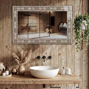 May include: A rectangular mirror with a decorative patterned border hangs above a wooden vanity with a white vessel sink. The mirror reflects a modern bathroom with a glass shower and wooden walls. A hanging plant adds a touch of greenery.