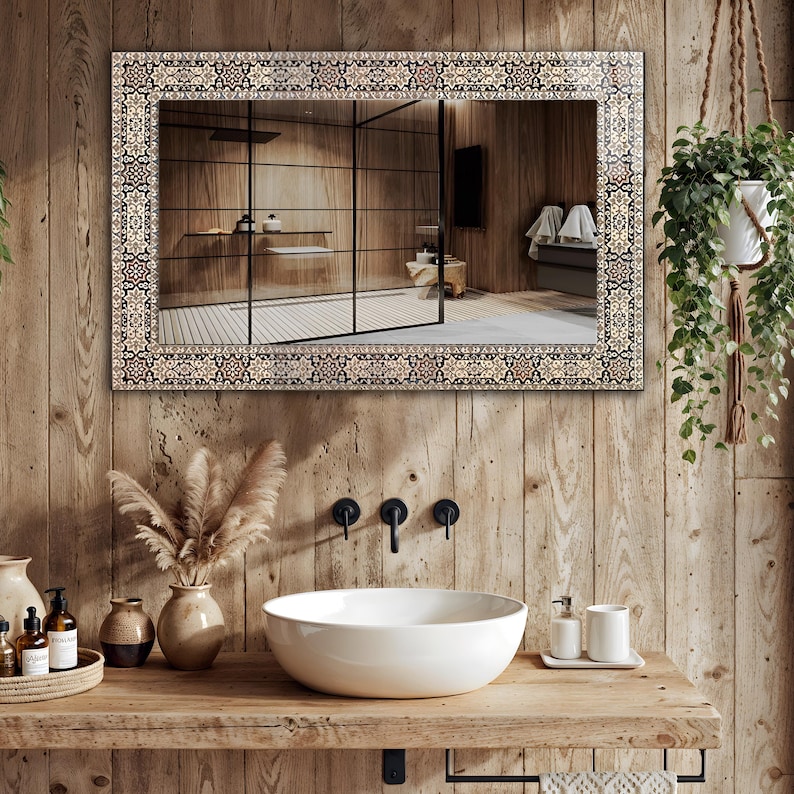 May include: A rectangular mirror with a decorative patterned border hangs above a wooden vanity with a white vessel sink. The mirror reflects a modern bathroom with a glass shower and wooden walls. A hanging plant adds a touch of greenery.