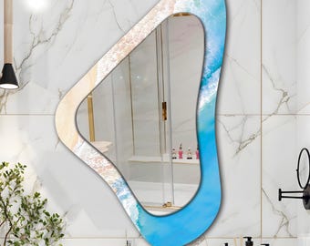 May include: An abstract-shaped wall mirror with a beach-themed design. The mirror's frame features a blue and white ocean wave pattern, with a sandy beach design on the opposite side. The mirror is mounted on a white marble tile wall.