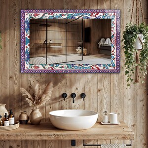 May include: Rectangular mirror with a decorative border featuring a floral pattern in blue, red, and green on a white background. The mirror is mounted on a wooden wall above a white sink, enhancing the bathroom's aesthetic.