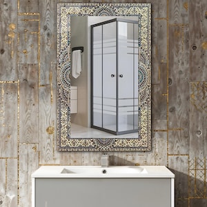 K&ouml;nnte beinhalten: Ein rechteckiger Spiegel mit einem verzierten, gemusterten Rahmen in Gold-, Blau- und Beiget&ouml;nen. Der Spiegel spiegelt eine moderne Duschkabine, ein wei&szlig;es Handtuch und einen grauen Waschtisch mit einem wei&szlig;en Waschbecken wider. Der Hintergrund ist eine Holzwand.