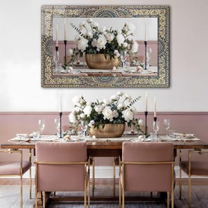K&ouml;nnte beinhalten: Ein gro&szlig;er rechteckiger Spiegel mit einem verzierten gold- und t&uuml;rkisfarbenen Rahmen h&auml;ngt &uuml;ber einem Esstisch. Der Tisch ist mit einem Blumengesteck in einer goldenen Schale, Kerzen und Gedecken dekoriert. Rosa St&uuml;hle mit goldenen Rahmen vervollst&auml;ndigen die Szene.