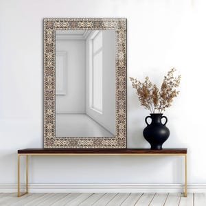 May include: A rectangular mirror with a decorative border featuring a traditional rug design in beige, black, and brown. The mirror is mounted above a dark wood console table with gold-colored legs. A black vase with dried plants sits on the table.