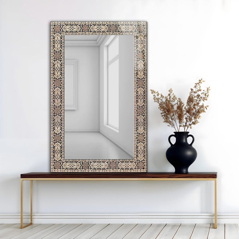 May include: A rectangular mirror with a decorative border featuring a traditional rug design in beige, black, and brown. The mirror is mounted above a dark wood console table with gold-colored legs. A black vase with dried plants sits on the table.