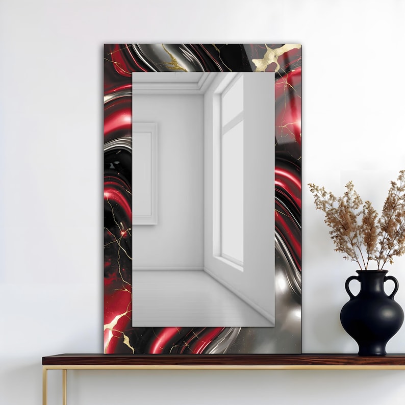 May include: Rectangular wall mirror with a decorative frame. The frame features a swirling pattern of red, black, silver, and gold. The mirror is displayed on a dark brown shelf with a black vase and dried plants.