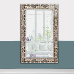 May include: Rectangular wall mirror with a decorative border featuring a repeating pattern in shades of beige, black, and brown. The mirror reflects a room with a white sofa, pillows, and a window with natural light.