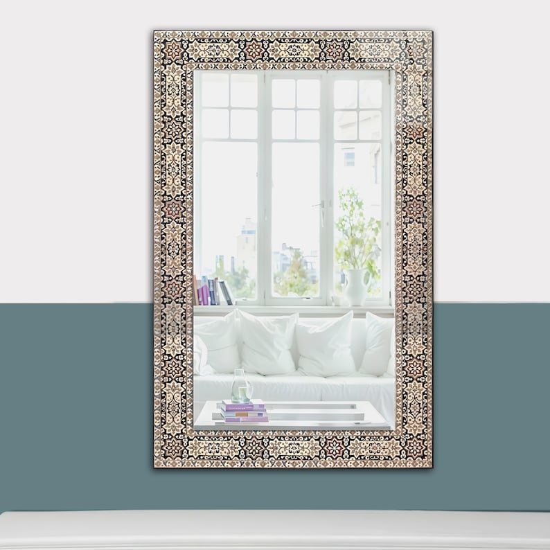 May include: Rectangular wall mirror with a decorative border featuring a repeating pattern in shades of beige, black, and brown. The mirror reflects a room with a white sofa, pillows, and a window with natural light.