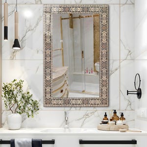 May include: Rectangular mirror with an ornate, patterned border in beige, black, and gold tones. The mirror is mounted on a white marble wall above a white sink. The reflection shows a bathroom with a shower and vanity.