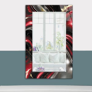 May include: Rectangular wall mirror with a vibrant, abstract design. The frame features a swirling pattern of red, black, and silver with gold accents. The mirror reflects a bright window and a white sofa with pillows.